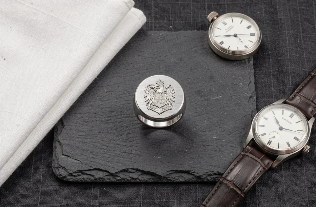 Chevalière homme en argent avec gravure héraldique posée sur ardoise dans une composition flat lay lifestyle masculine